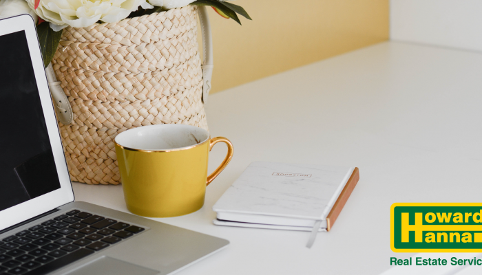 a yellow mug and notebook sit by a laptop on a desk as a howard hanna agent trains offscreen